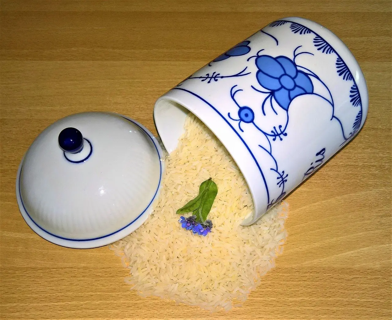 Blue and white ceramic jar spilled rice and a small flower on a wooden surface.